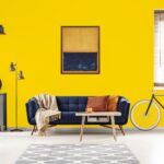 Yellow and blue painting hanging on white wall in bright living room interior with grey cupboard, gold lamp, sofa with blanket and pillows and bike standing under window with blinds