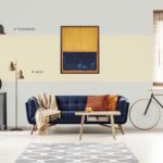 Yellow and blue painting hanging on white wall in bright living room interior with grey cupboard, gold lamp, sofa with blanket and pillows and bike standing under window with blinds