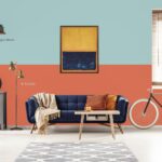 Yellow and blue painting hanging on white wall in bright living room interior with grey cupboard, gold lamp, sofa with blanket and pillows and bike standing under window with blinds